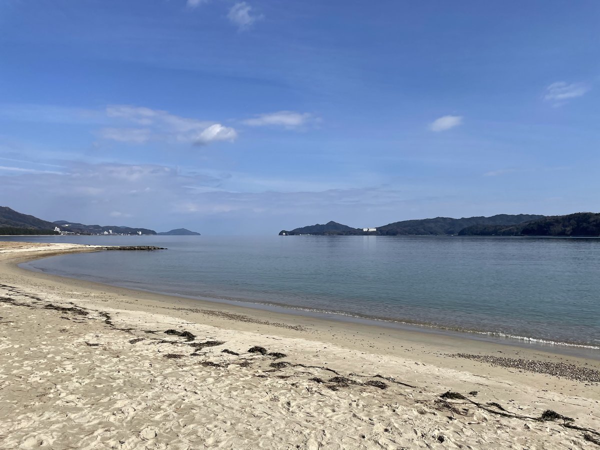A wide sandy beach and calm blue sea under a blue sky, with low green hills across the water