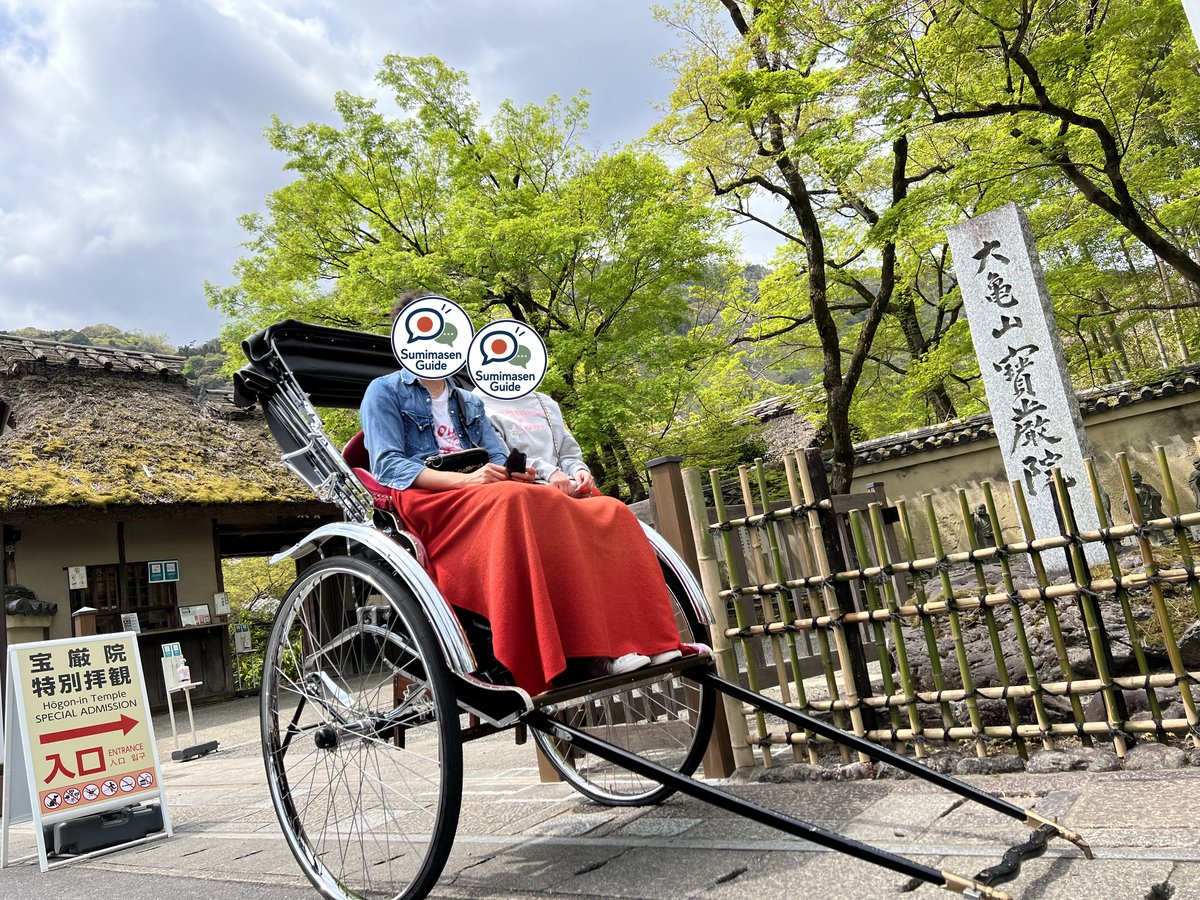 Two people sitting in a black rickshaw with a red blanket in front of a temple gate and a stone marker, under trees with bright green young maple leaves; their faces are covered with the Sumimasen Guide logo