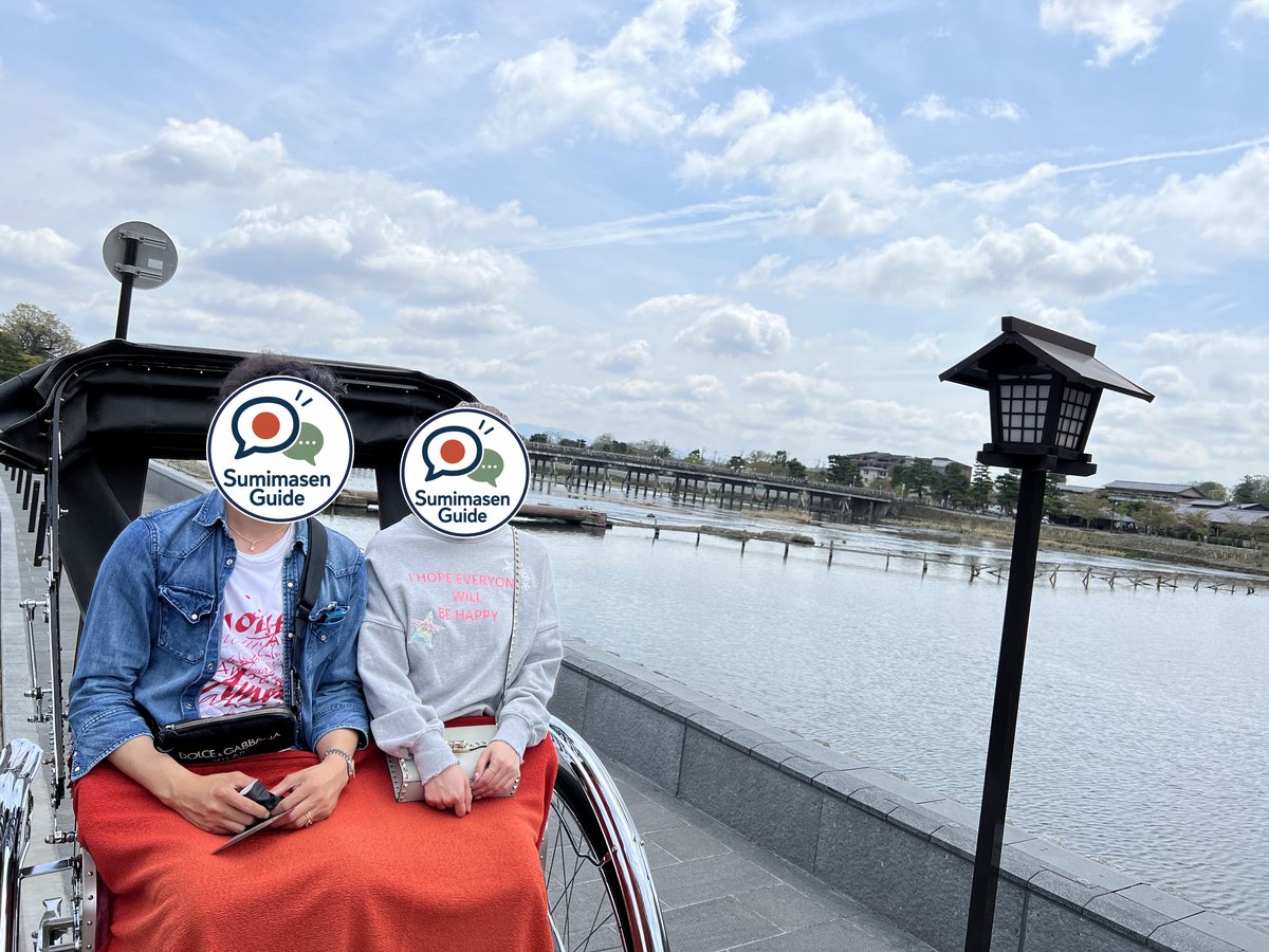 Two people sitting in a rickshaw on a riverside path with a lantern, with a long bridge across the river behind them; their faces are covered with the Sumimasen Guide logo