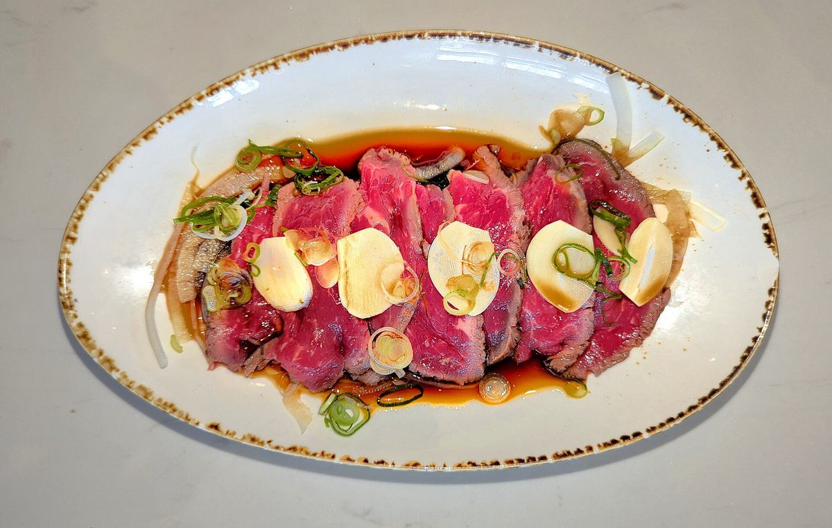 Slices of beef tataki, seared at the edge and red in the middle, with thin slices of garlic, onion and green onion in a dark sauce