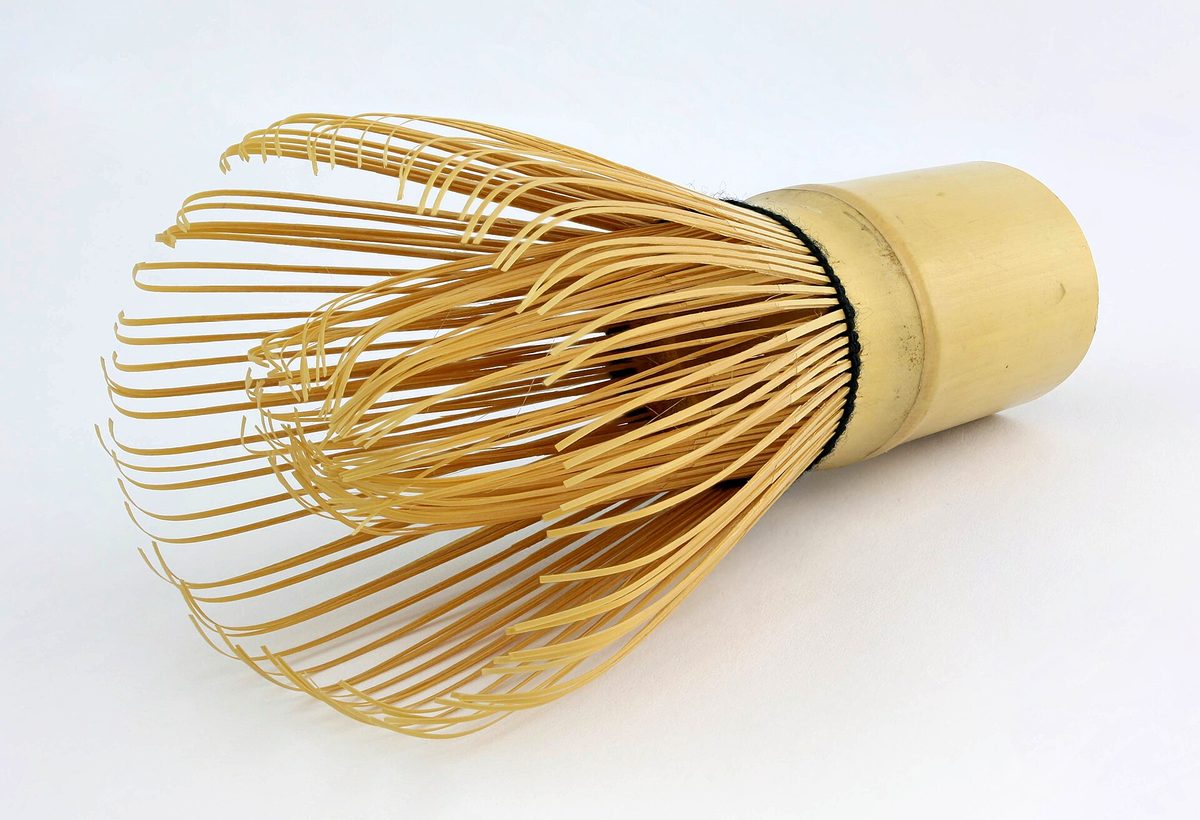 A bamboo tea whisk lying on its side on a white background, its many fine curved tines spreading out