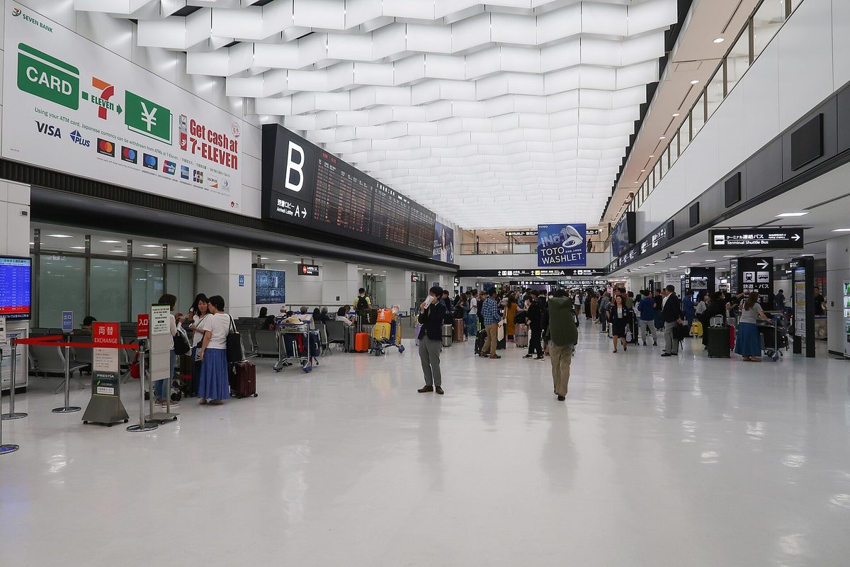 캐리어를 끄는 여행자들과 왼쪽에 카드·환전 안내판이 보이는 밝은 나리타 공항 제2터미널 도착 로비
