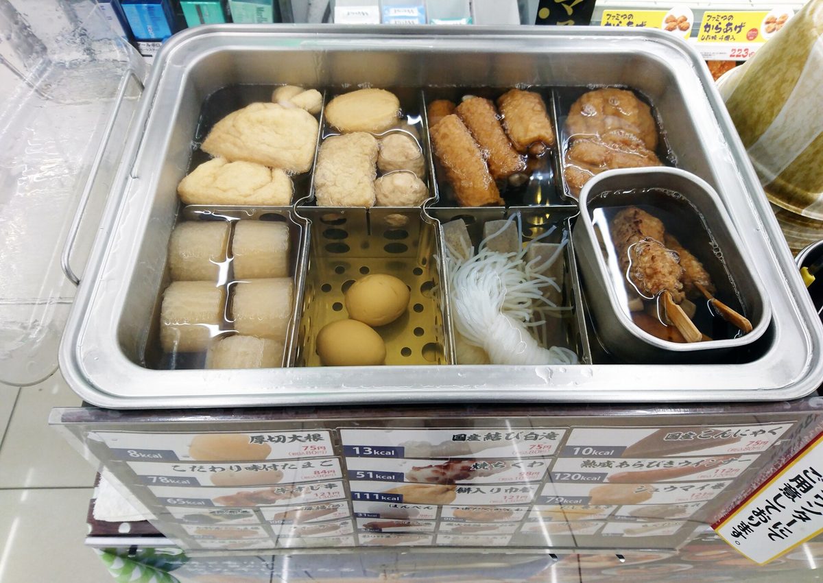 A metal oden pot at a convenience store counter in Japan, with daikon, eggs, shirataki noodles, fried tofu and skewered beef tendon simmering in separate sections