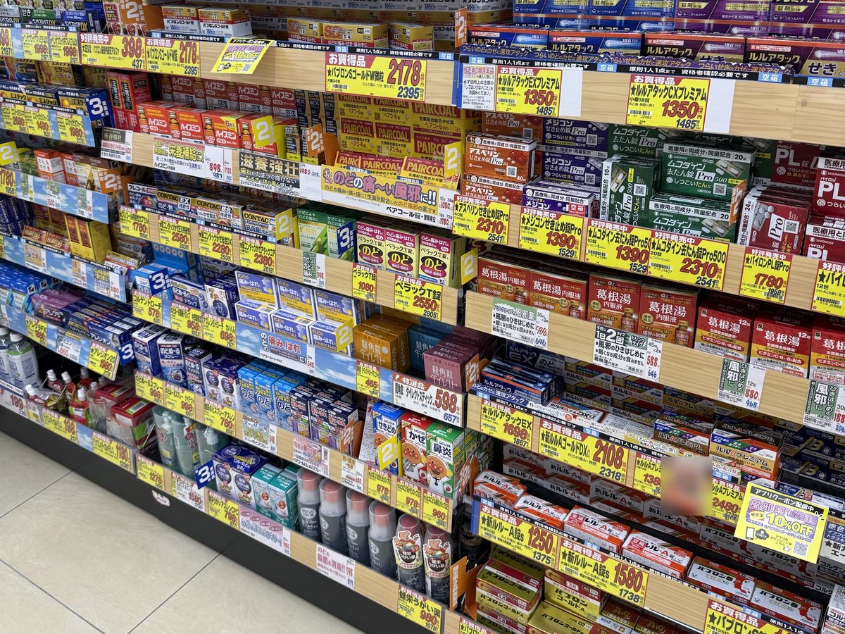Shelves of cold medicine boxes with yellow price tags along a drugstore aisle, with a 'one per person' sign on the top shelf