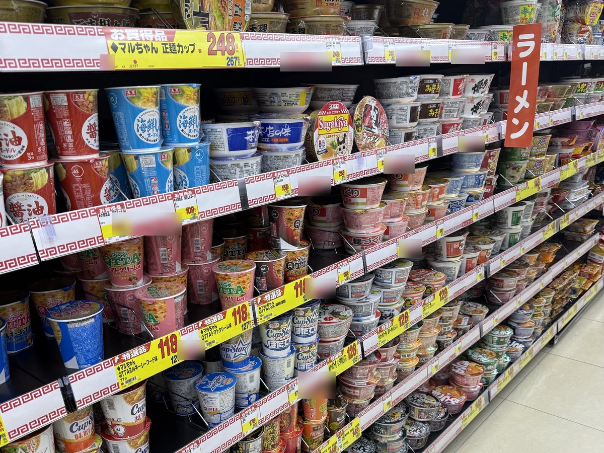 A long aisle of cup noodles on shelves with red and white price rails and a red 'ramen' sign