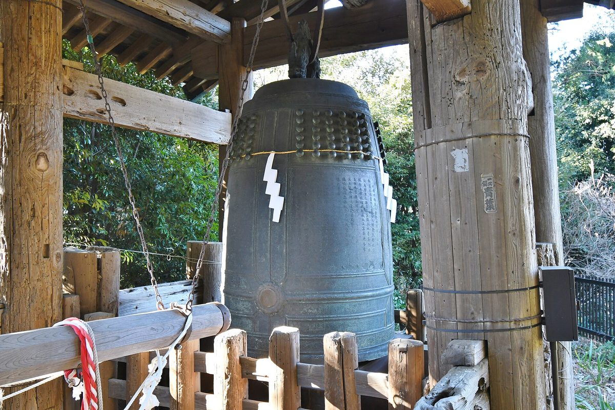 A large dark bronze temple bell with rows of round knobs, hanging under a wooden roof among trees, with a white paper streamer on it