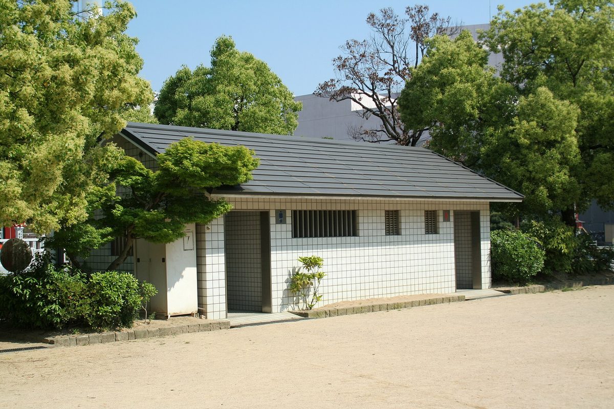 A small single-story public toilet building with a gray roof among trees in a park