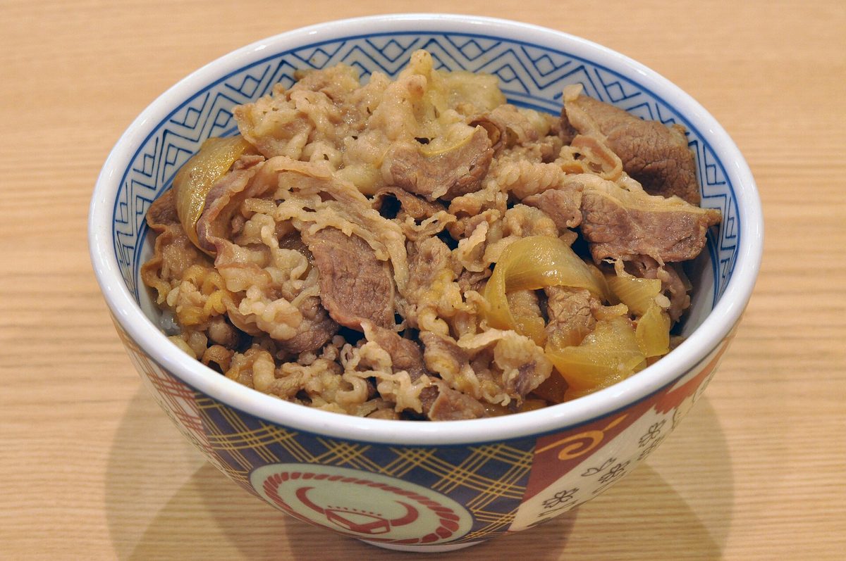 A bowl of gyudon: thinly sliced simmered beef and onion over rice, in a patterned bowl on a wooden table