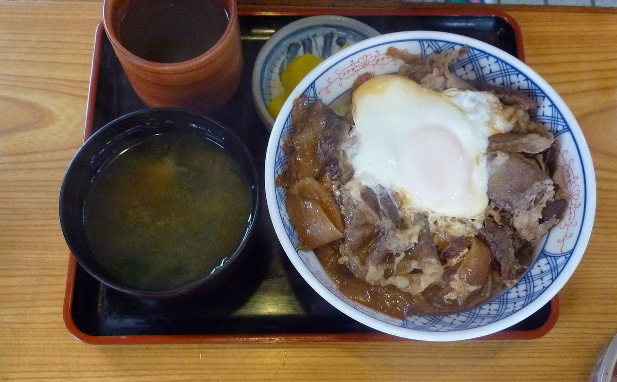 A tray with a beef bowl topped with a soft-cooked egg, a bowl of miso soup, yellow pickles and a cup of tea