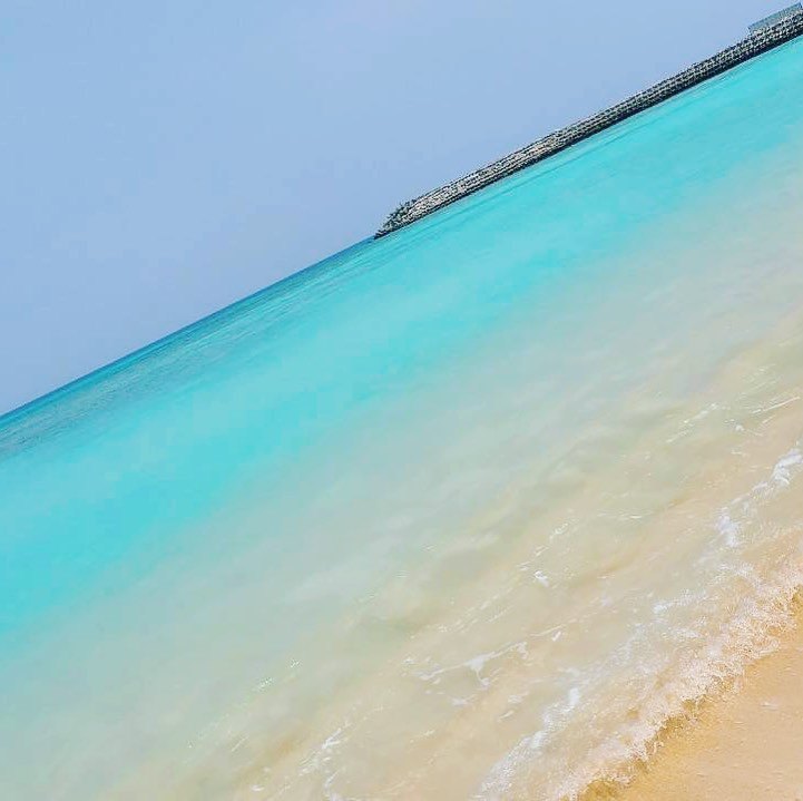 Clear turquoise water lapping on a pale sand beach, with a stone breakwater far out on the sea