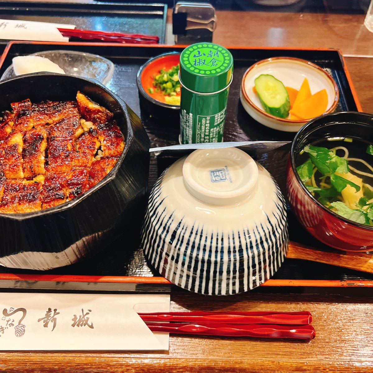 Hitsumabushi in a black wooden tub on a tray, with an upside-down rice bowl, a small dish of green onion and wasabi, a green can of sansho pepper, pickles and a clear soup