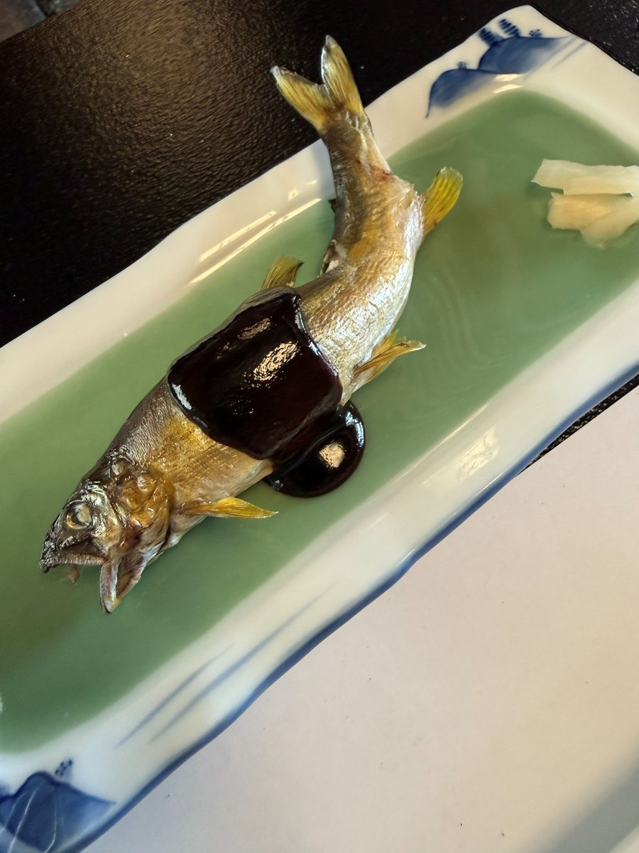 A whole grilled ayu topped with glossy dark miso on a long green and blue plate, with pickled ginger on the side