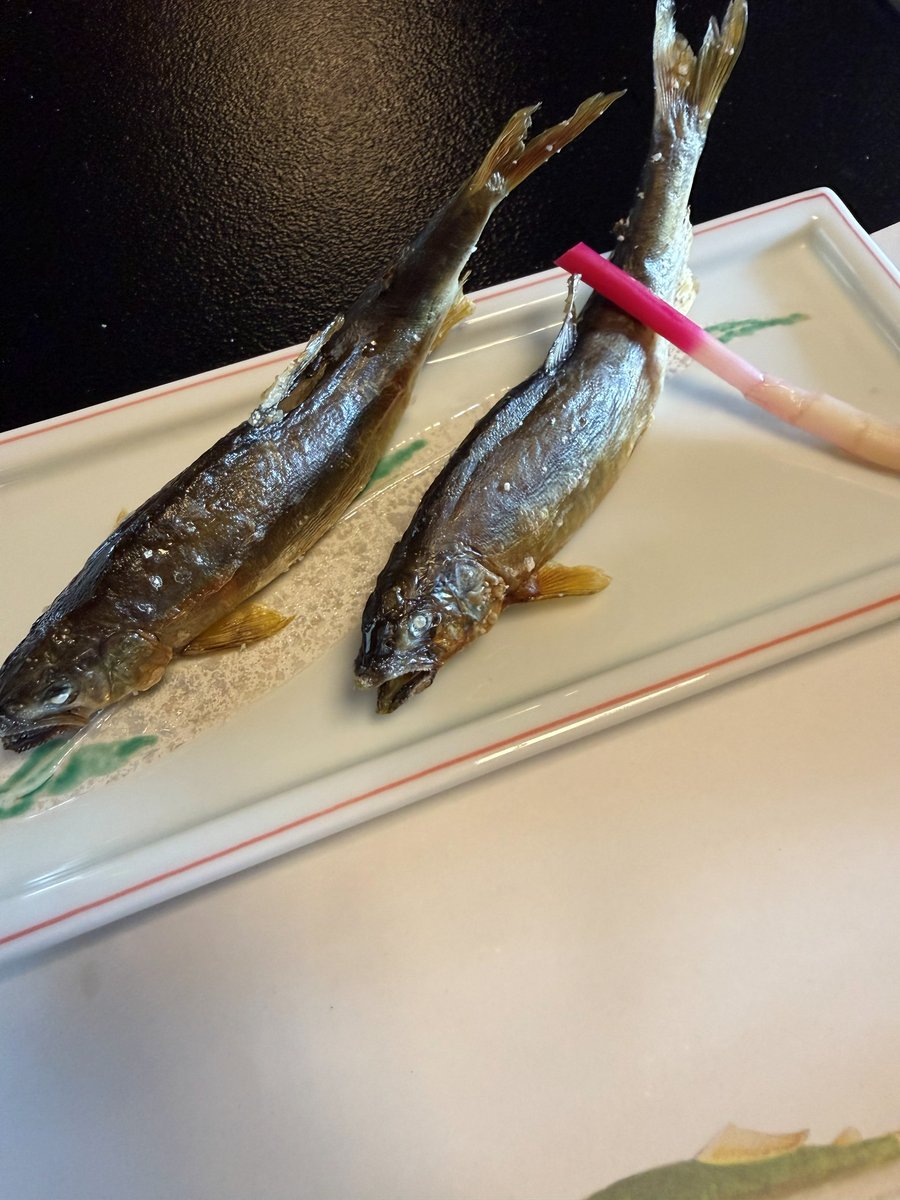Two whole grilled ayu with salt on their skin, side by side on a long white plate with a pink pickled ginger shoot