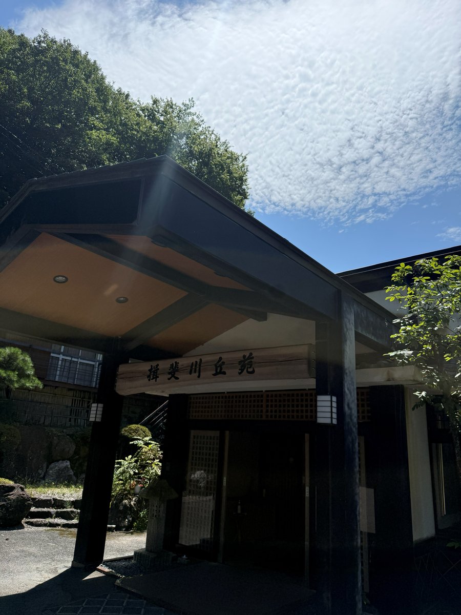 The entrance of a Japanese inn under a wide wooden roof, with a long wooden sign reading 揖斐川丘苑 and trees around it