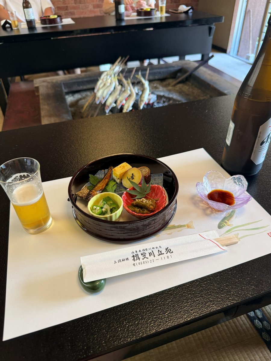 A black table with a lacquered tray of small appetizers, a glass of beer and chopsticks in a paper sleeve; behind it, ayu on skewers stand around the charcoal of a sunken hearth