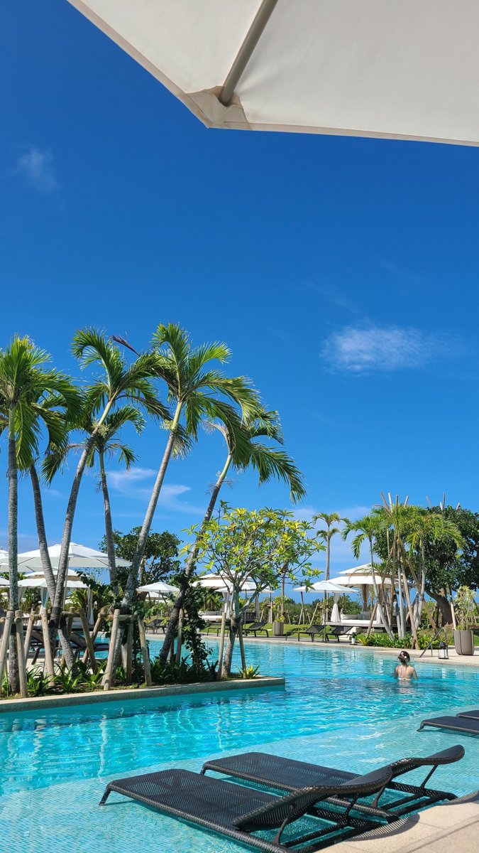 A long bright blue outdoor pool with sun loungers in the water, tall palm trees and white parasols under a clear sky