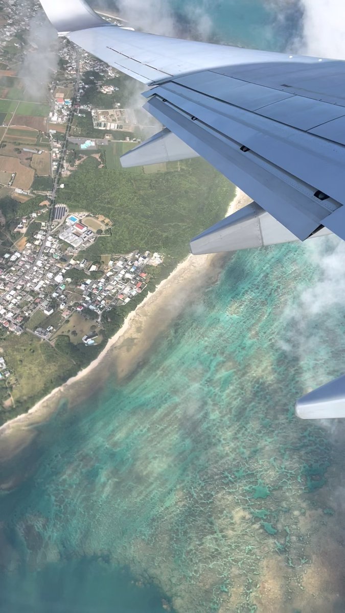 A view from a plane window of a coastline with houses, green fields and wide shallow turquoise water, with the plane's wing above