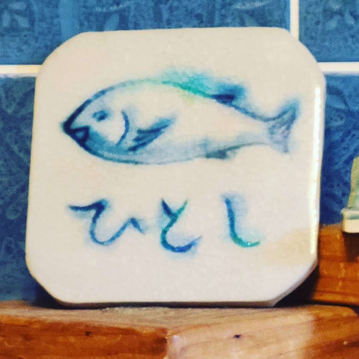 A small square ceramic plate painted with a blue fish and the name Hitoshi in Japanese letters, standing in front of blue tiles
