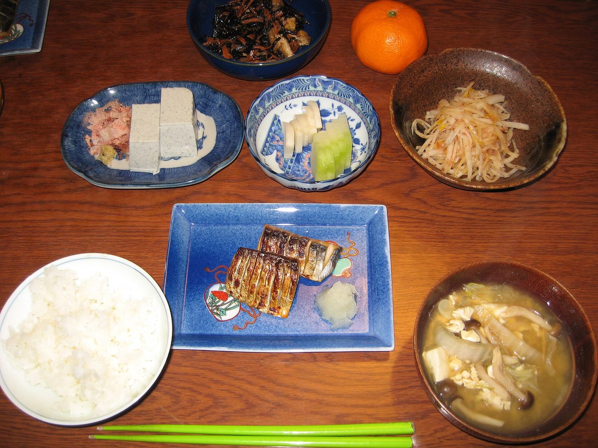 A home dinner on a wooden table: a bowl of white rice, miso soup with tofu and mushrooms, grilled fish and small side dishes