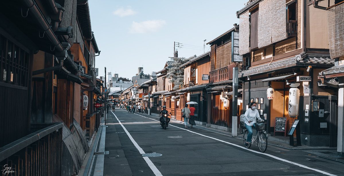 A person riding a bicycle on the left side of a quiet street lined with wooden houses