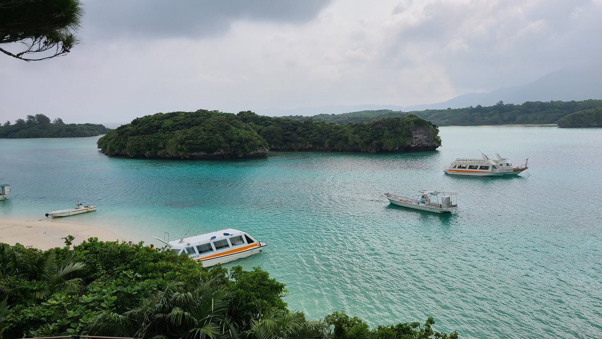Looking down through trees at turquoise water, a white beach, glass-bottom boats and a small green island in Kabira Bay