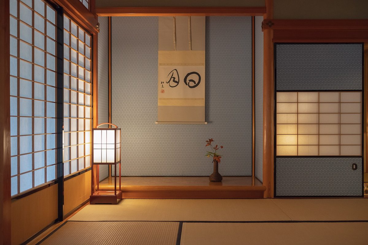 A quiet tatami room with paper sliding doors, a paper lantern and an alcove holding a hanging scroll and a small flower arrangement