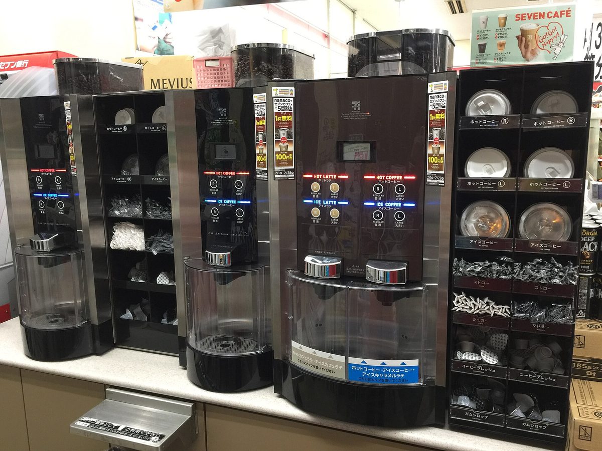 Three self-service coffee machines on a counter at a 7-Eleven in Japan, with lids, straws, sugar and stirrers on a black shelf to the right