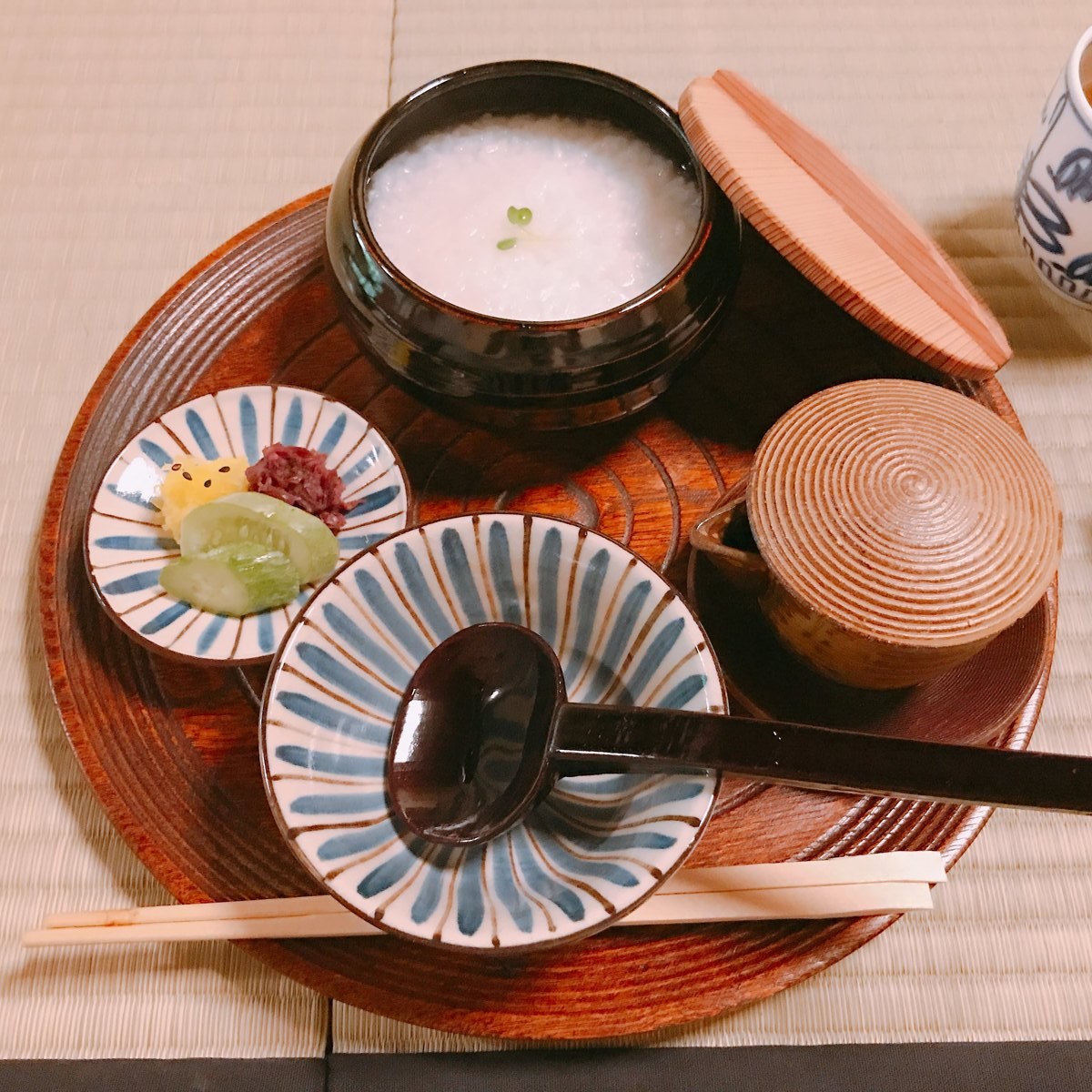 A lacquered round tray seen from above, with a bowl of white rice porridge, a black lidded bowl, a ladle and small patterned dishes on a tatami mat