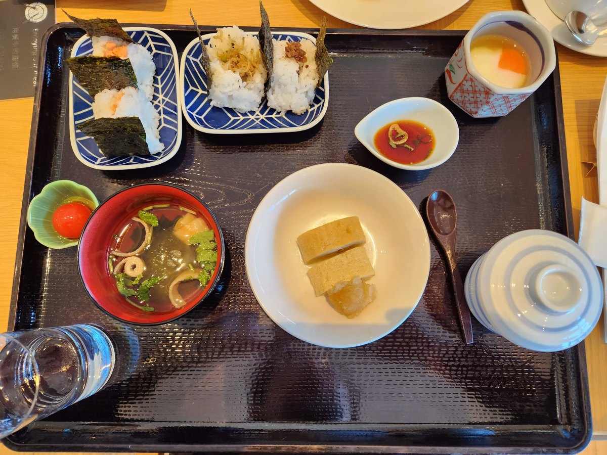 A black tray with rice balls wrapped in seaweed, a rolled omelet, a bowl of soup, a small tomato and small dishes
