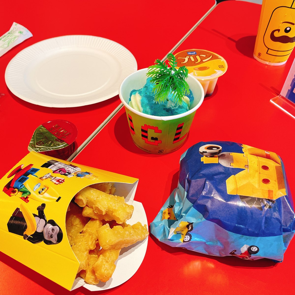 A children's meal on a red table: fries in a LEGO-printed box, a blue drink in a cup and a wrapped LEGO-themed item