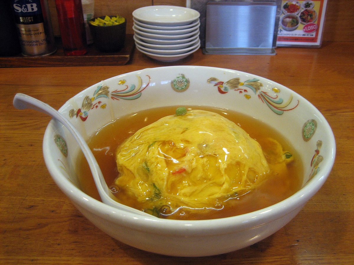 A bowl of tenshinhan: a fluffy omelet on rice covered in glossy brown sauce, on a restaurant counter