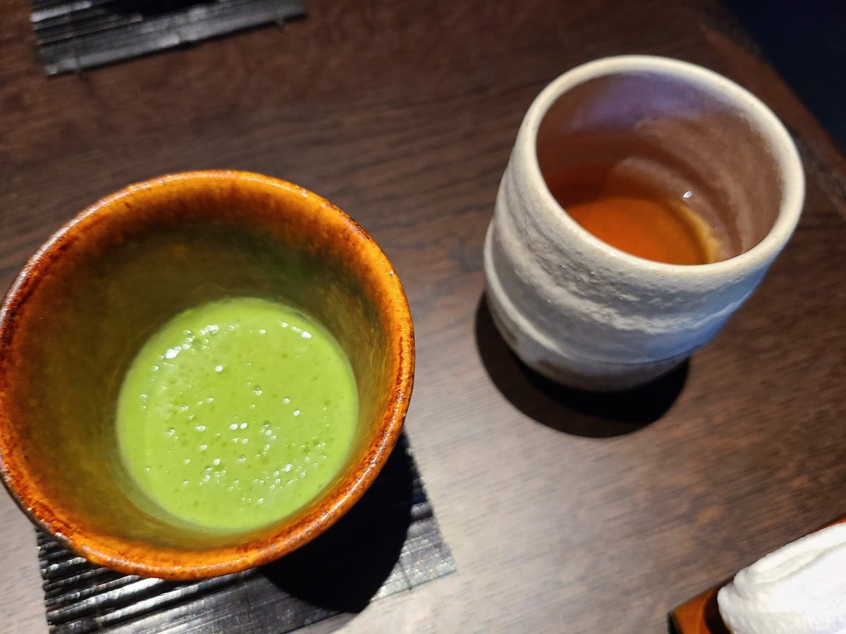 A bowl of bright green matcha next to a cup of brown tea on a dark table