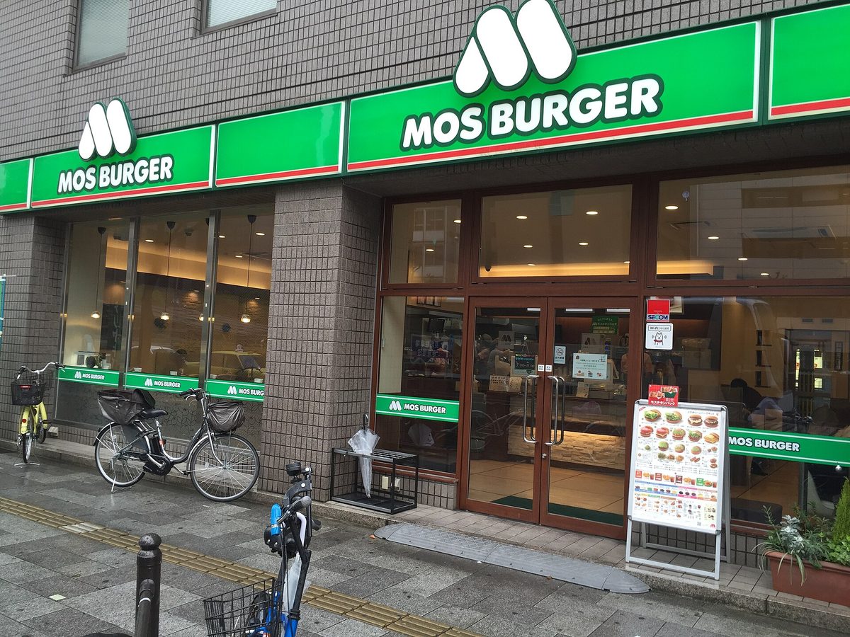 초록색 간판과 큰 유리창이 있는 모스버거 매장 앞. 밖에는 자전거가 세워져 있고 문 옆에 메뉴판이 있습니다