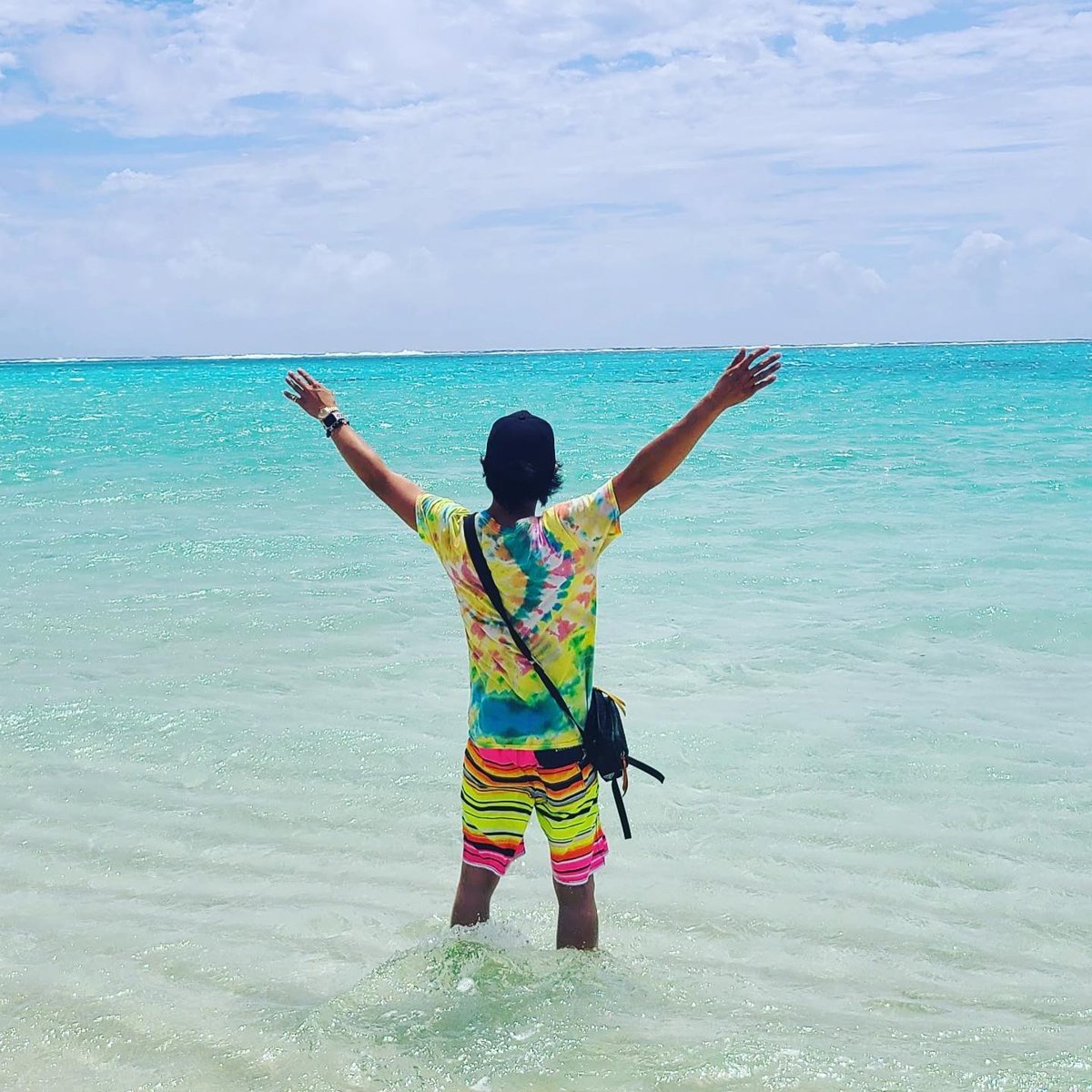 A person seen from behind with both arms raised, standing in shallow turquoise sea under a sky of white clouds