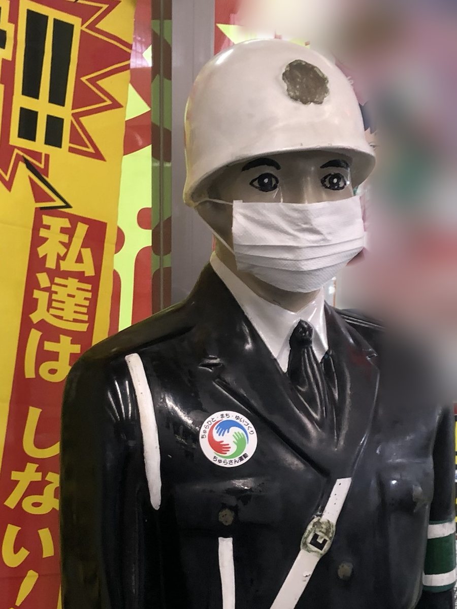Mamoru-kun, a police officer doll in a white helmet and a face mask, standing beside traffic-safety banners