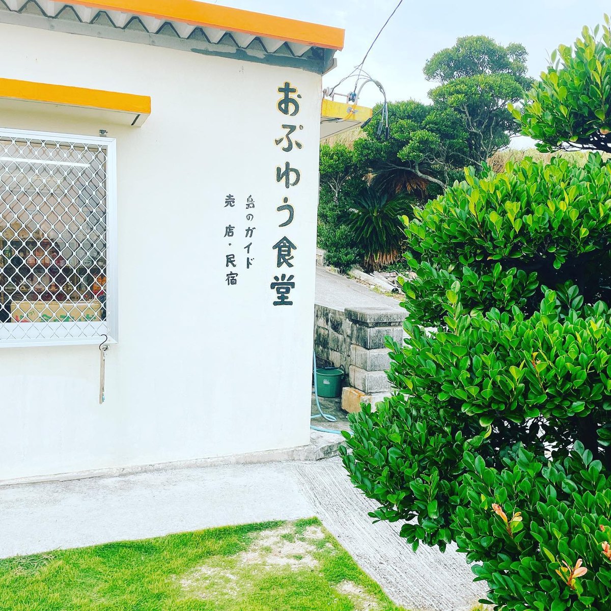 A white building with an orange roof edge and Japanese lettering on the wall, beside green trees and a lawn