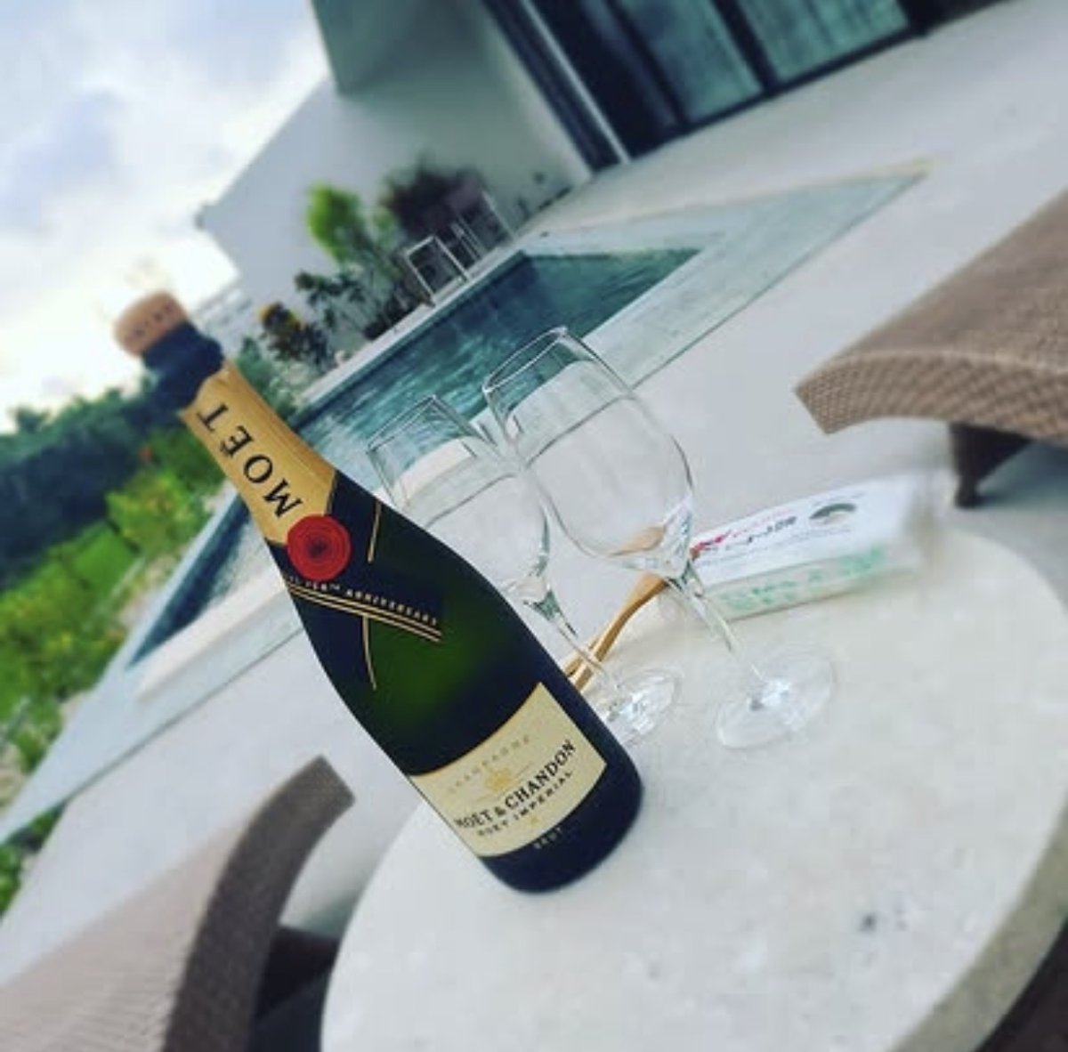 A bottle of champagne and two glasses on a round white table, with a private pool and a white villa behind