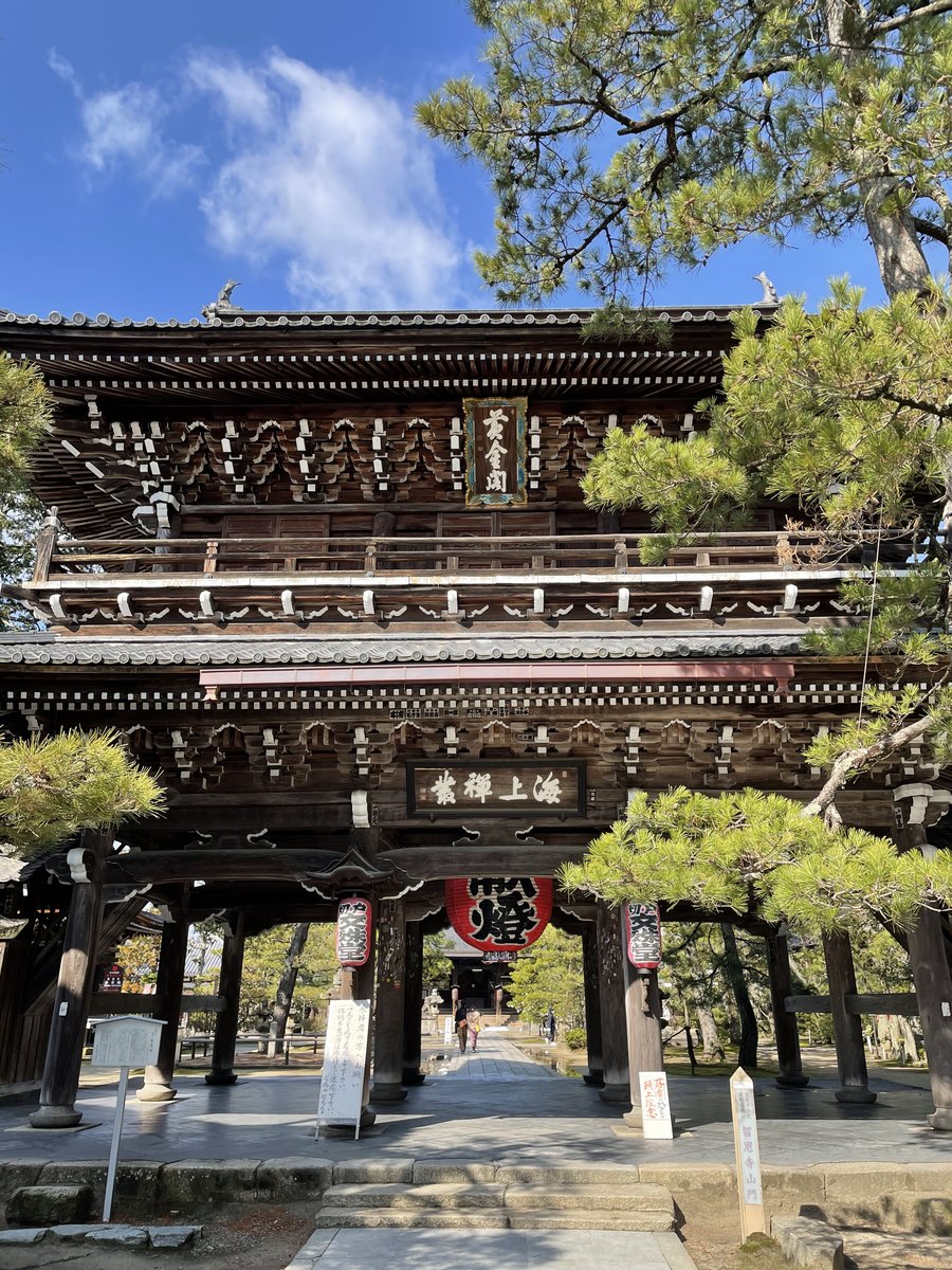 A large two-story wooden temple gate with tiled roofs and lanterns, framed by pine trees under a blue sky