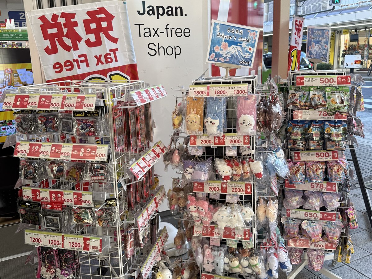 드러그스토어 앞 'Tax-free Shop' 간판 아래, 캐릭터 인형 마스코트와 일본 키홀더·자석이 걸린 회전 진열대