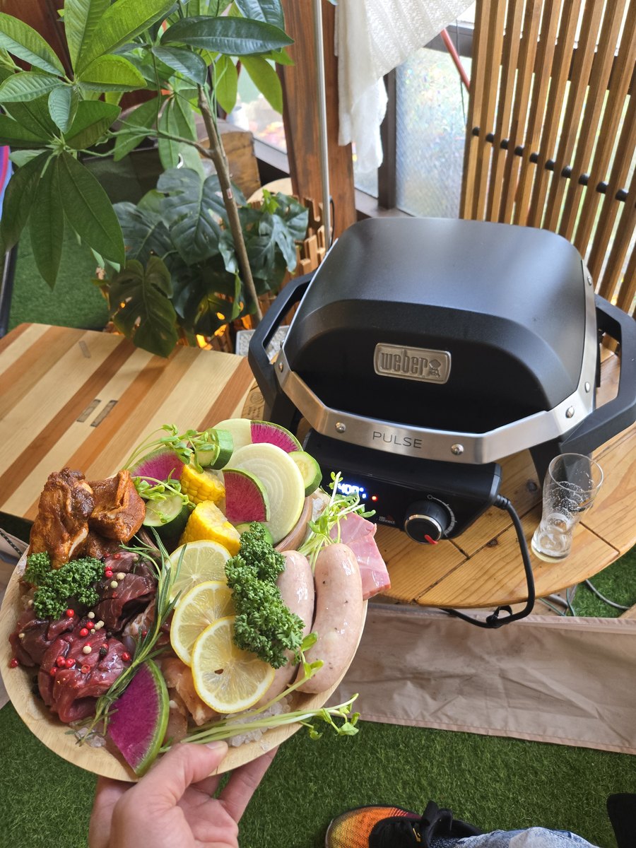 A hand holding a plate of seasoned meat, sausages, lemon slices and colorful vegetables in front of an electric grill
