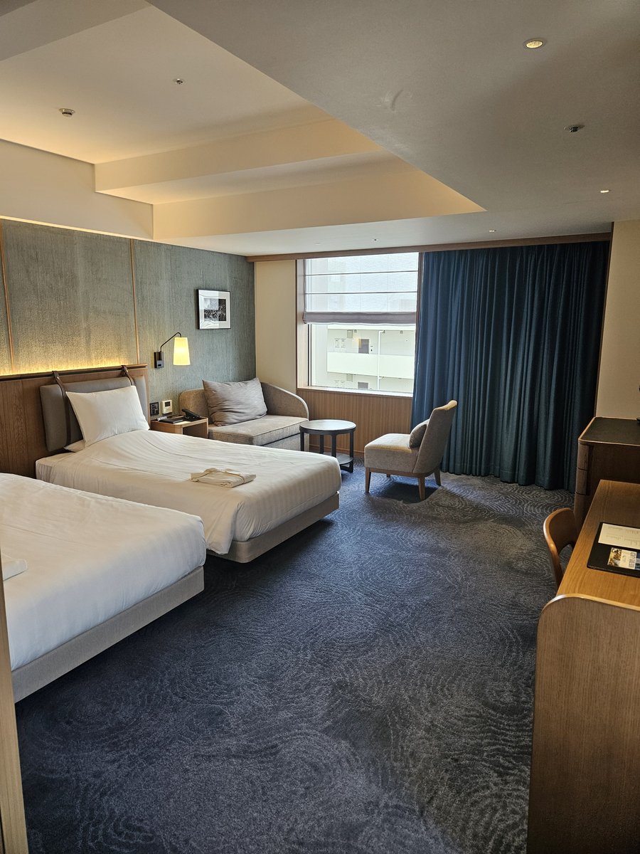 A corner hotel room with two white beds, a small sofa and armchair by a window with navy curtains, soft indirect lighting and a patterned blue carpet