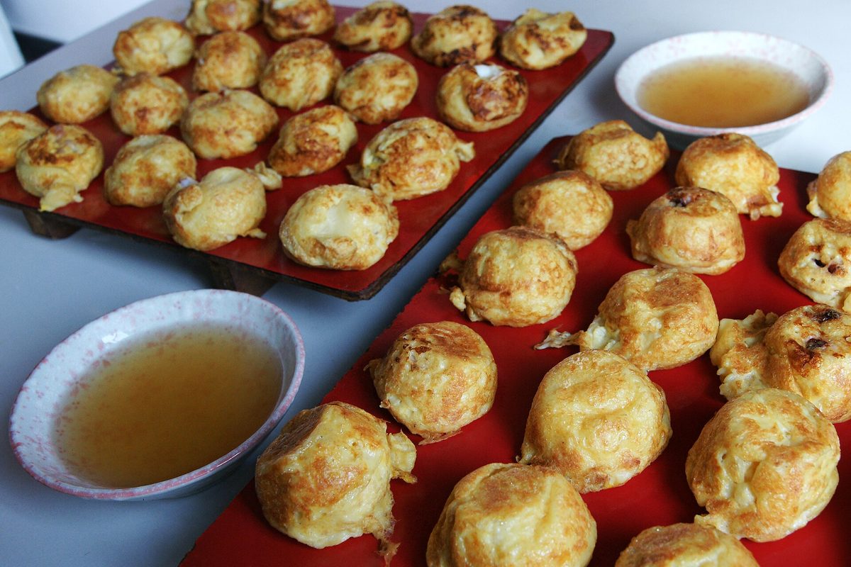 Soft, egg-yellow Akashi-yaki dumplings lined up on a red board, with small bowls of dashi for dipping