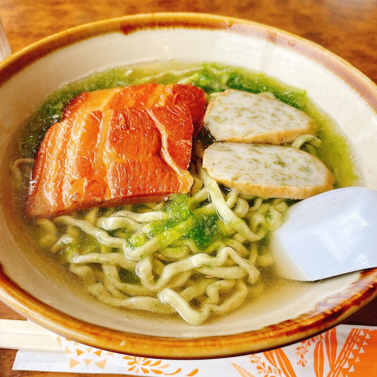 A bowl of Okinawa soba with pale green noodles, a slab of braised pork, two slices of fish cake and green sea lettuce in a clear broth