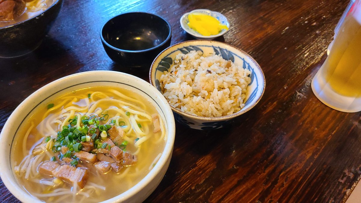 A bowl of Okinawa soba with thick noodles, pork and green onion, next to a bowl of mixed rice and a small bowl of soup on a wooden table