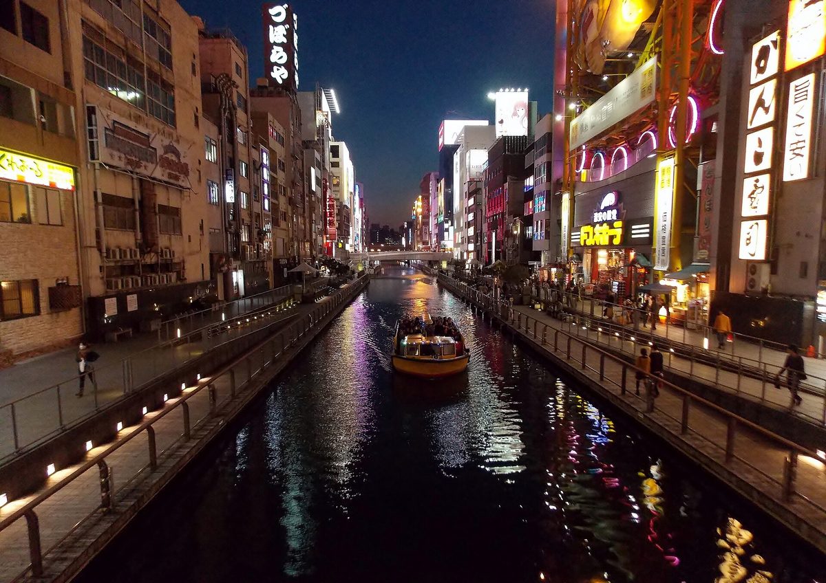 The Dotonbori canal in Osaka at night, with lit-up signs and restaurants on both banks and a tour boat on the water