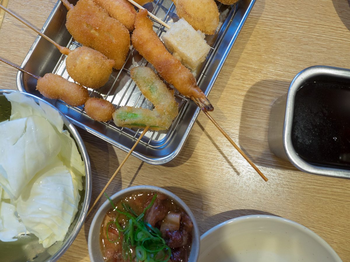 Kushikatsu skewers on a metal tray next to a sauce pot and a bowl of raw cabbage