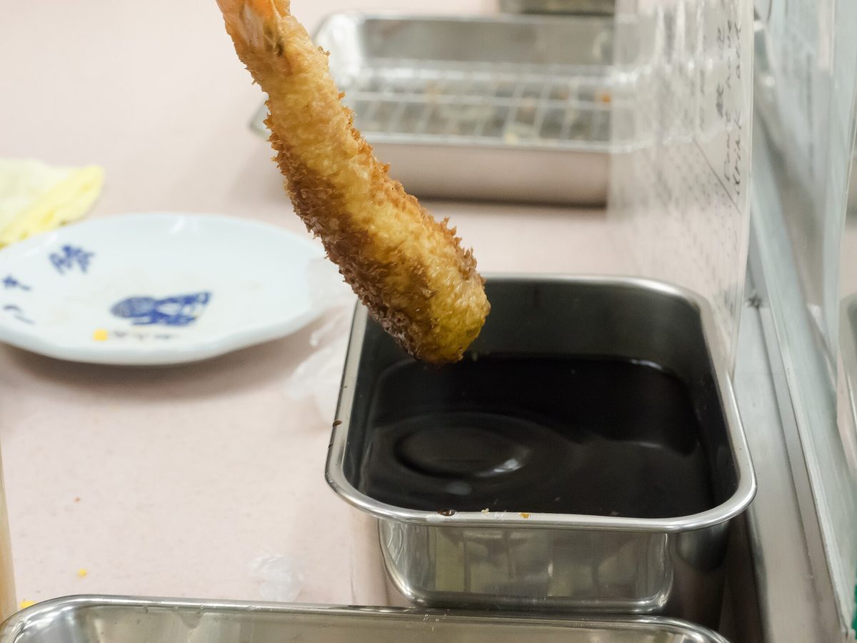 A fried shrimp skewer being dipped into a metal tray of sauce