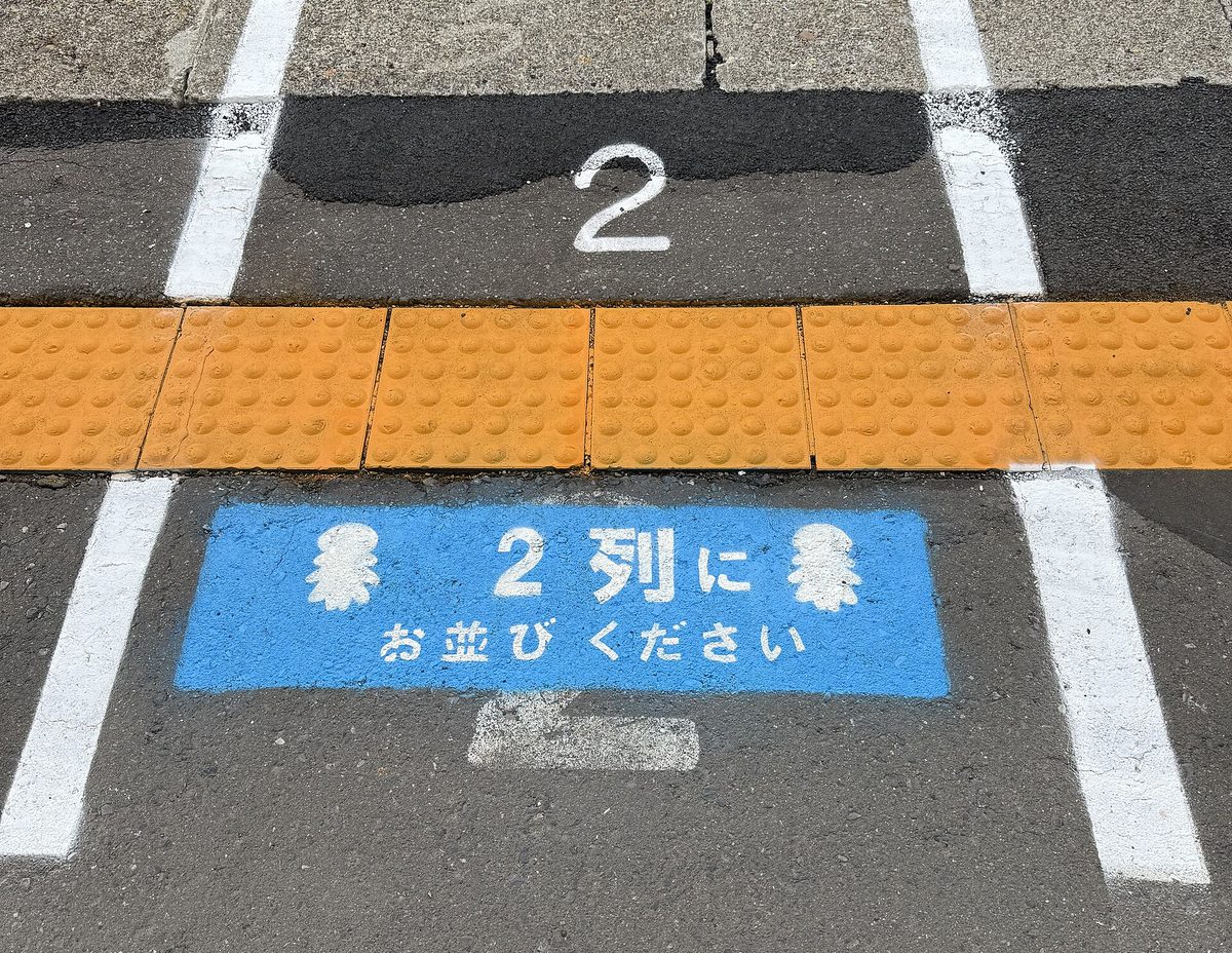 Painted lines and a number 2 on a station platform floor, with a blue sign asking people to line up in two rows