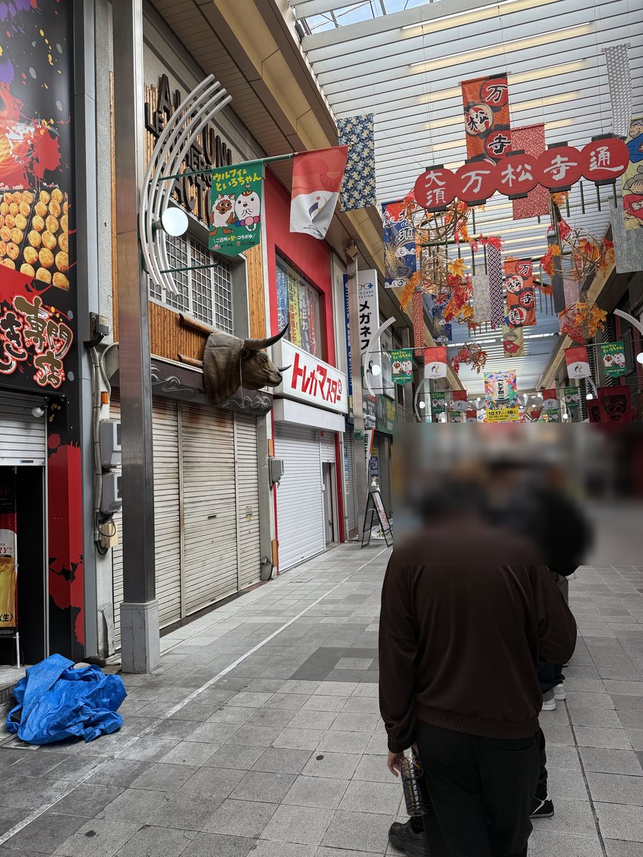 People standing in a line in a covered shopping arcade in front of closed shop shutters, with their heads blurred