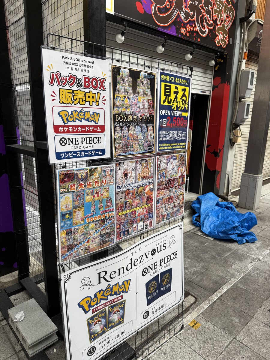 A sign board outside a card shop with posters for Pokémon Card Game and ONE PIECE Card Game packs and boxes, and oripa at 500 yen and 2,500 yen